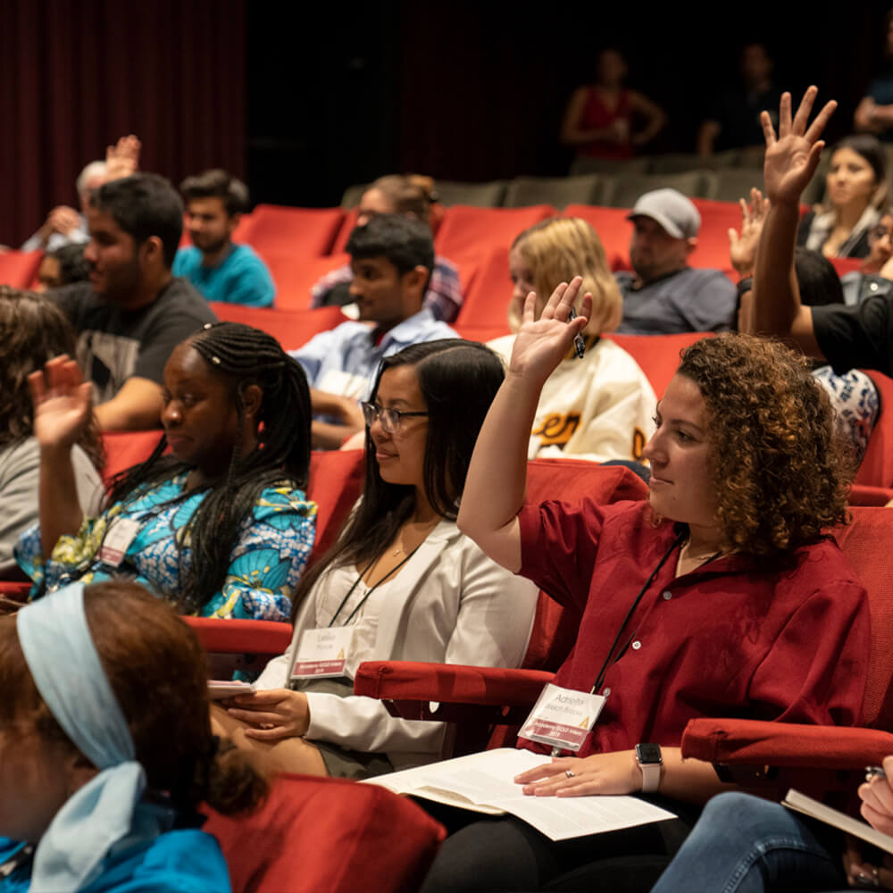 students sit in theater seats with notepads and raise their hands while looking at the presenter